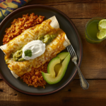 A plate of green chile enchiladas served with Mexican rice, refried beans, avocado slices, and a dollop of sour cream.