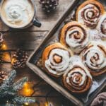 Warm cinnamon rolls topped with cream cheese frosting, placed on a rustic table with holiday decor.