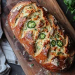 Freshly baked vegan jalapeño cheese artisan bread loaf with a golden crust, topped with melted vegan cheese and jalapeño slices on a cutting board.