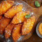 Golden fried sweet potato turnovers with a flaky crust, dusted with powdered sugar, on a rustic wooden board with vanilla glaze and mint leaves.