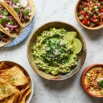 Cinco de Mayo party food spread with guacamole, tacos, and street corn dip on a marble surface